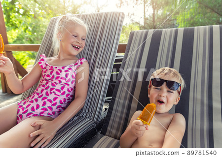 Two cute adorable caucasian blond little siblings enjoy having fun relaxing and eat fruit popsicle icecream sitting on sunbed in yard garden near pool sea beach. Brother and sister chilling outdoor 89781540