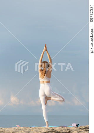 Girl in white sporty clothes practicing yoga, tree pose on the beach. Vrikshasana. Young woman doing yoga with the sea behind. 89782584