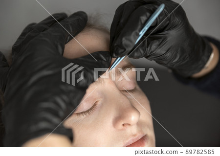 Procedure of eyebrow correction, young woman in beauty salon. Cosmetologist in black gloves plucks eyebrows with tweezers. 89782585