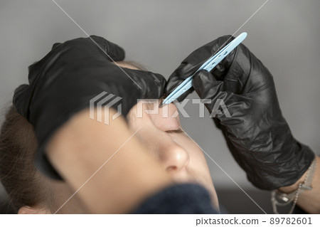 Young woman on the procedure of eyebrow correction. Cosmetologist in black gloves plucks eyebrows with tweezers. 89782601