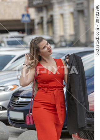 Young sexy woman with curvy body shape standing on street and car background wearing red close fitting sleeveless dress with deep neckline. 89782996