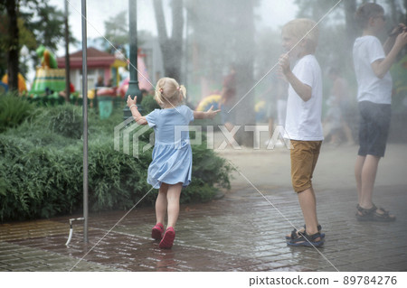 Children play under the water jets in summer park. Breeze on hot day. Playground. Children play under the water jets in summer park. Breeze on hot day. Playground. 89784276