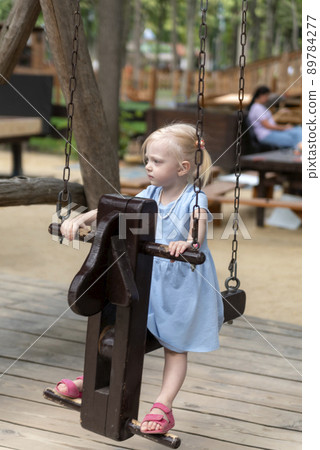 Little blonde girl in blue dress is swinging on wooden swing in summer park outdoors. Vertical frame. 89784277