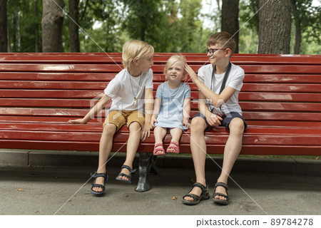 Two brothers and little sister are resting in park on summer day. Portrait of siblings sit on bench. Two brothers and little sister are resting in park on summer day. Portrait of siblings sit on bench. 89784278