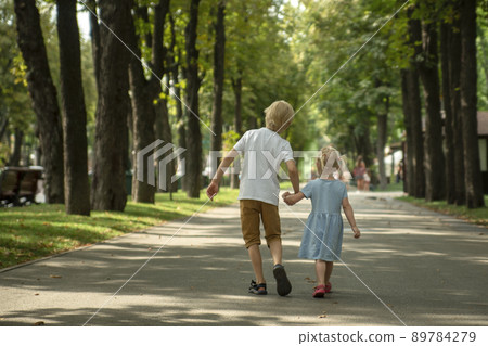 Little brother and sister walk in park. Older brother holds his sister's hand and walks along alley of park. Back view. Little brother and sister walk in park. Older brother holds his sister's hand and walks along alley of park. Back view. 89784279