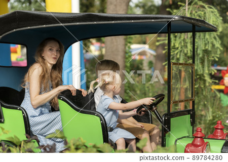 Two young drivers of the child roll their mother in car in an amusement park. Nanny looks after the children in theme park. 89784280