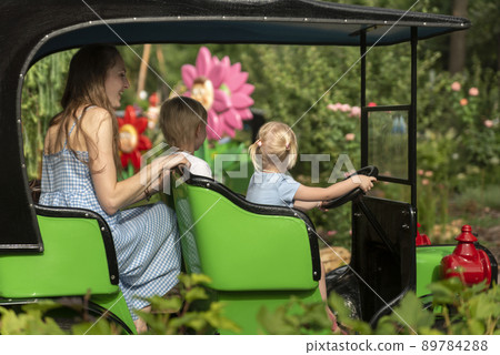 Two young child and their mother in little car in an amusement park. Nanny looks after the children in theme park. 89784288
