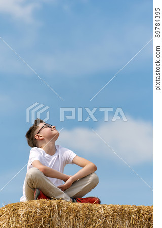 Happy teenage boy sits on haystack against blue sky. Child enjoys the fresh air and the sun in the village. Vertical frame Happy teenage boy sits on haystack against blue sky. Child enjoys the fresh air and the sun in the village. Vertical frame 89784395