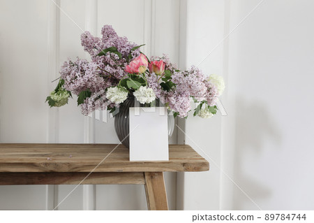Vintage floral stationery mockup. Blank greeting card, invitation. Vase with bouquet of lilac, viburnum and tulips on old wooden stool. White wooden wall background. Empty copy space. Vintage floral stationery mockup. Blank greeting card, invitation. Vase with bouquet of lilac, viburnum and tulips on old wooden stool. White wooden wall background. Empty copy space. 89784744