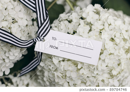 Closeup of blank retro gift tag, price label mock-up with striped black white ribbon. Blooming blurred Hydrangea blossoms. Birthday concept. Summer flowers. Top view. 89784746