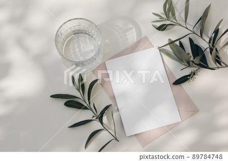 Modern summer wedding stationery still life. Drinking glass with shadows. Blank blush pink envelope, card mock up scene. White table background in sunlight. Green olive tree branches. Flat lay, top Modern summer wedding stationery still life. Drinking glass with shadows. Blank blush pink envelope, card mock up scene. White table background in sunlight. Green olive tree branches. Flat lay, top 89784748