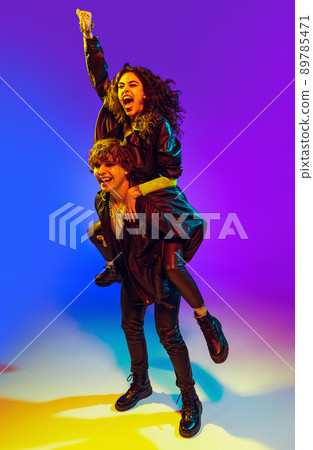 Young couple of rock-and-roll musicians wearing black leather outfits gesturing, shouting and posing on blue yellow background in neon light 89785471