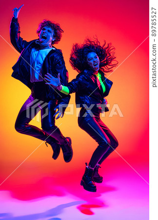 Excited young man and girl in black leather outfits having fun, gesturing, shouting and jumping on yellow-red background in neon light Excited young man and girl in black leather outfits having fun, gesturing, shouting and jumping on yellow-red background in neon light 89785527