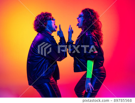 Excited young man and girl in black leather outfits having fun, gesturing, shouting and shaking head over yellow-red background in neon light Excited young man and girl in black leather outfits having fun, gesturing, shouting and shaking head over yellow-red background in neon light 89785568