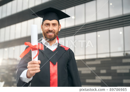 Front view of male student graduating from high school. Attractive young male wearing mortarboard and graduate gown, standing, showing diploma, smiling. Concept of success. 89786985