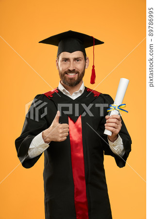 Front view of male with beard graduating from college, university. Handsome student standing, holding diploma, showing super, smiling. Isolated on yellow studio background. Front view of male with beard graduating from college, university. Handsome student standing, holding diploma, showing super, smiling. Isolated on yellow studio background. 89786995