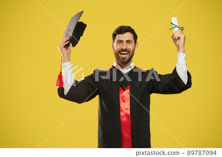 Front view of happy student holding diploma and mortarboard. Master, bachelor standing, smiling, looking at camera, wearing graduate gown, cheerful. Isolated on yellow background. 89787094
