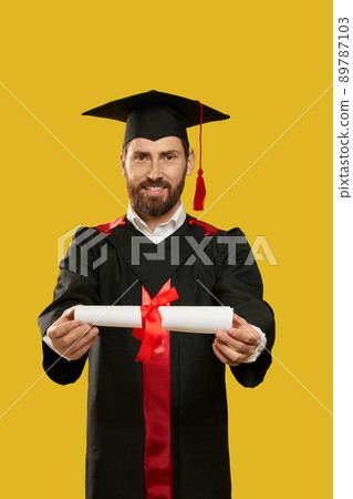 Front view of happy student getting bachelor, master degree. Young man with beard standing, holding, showing, giving diploma, smiling. Isolated on yellow studio background, 89787103