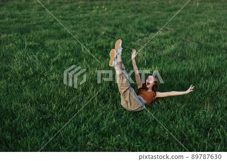 A young woman playing games in the park on the green grass spreading her arms and legs in different directions falling and smiling in the summer sunlight A young woman playing games in the park on the green grass spreading her arms and legs in different directions falling and smiling in the summer sunlight 89787630