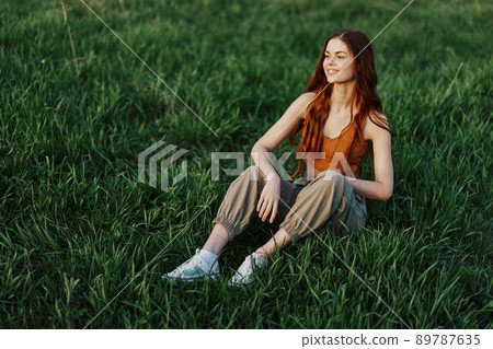 A beautiful skinny woman sits on the green grass outdoors in casual comfortable clothes and relaxes from exercising in the sunset light 89787635