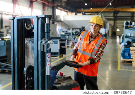 Young female engineer driving electric forklift while moving along warehouse 89788647