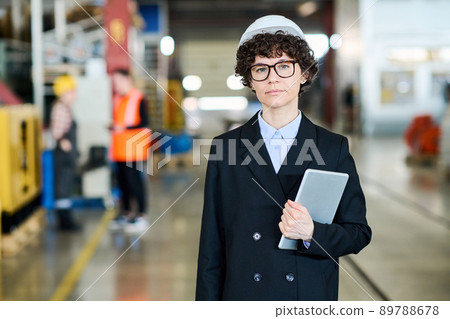 Young female factory worker in black formalwear and hardhat holding tablet Young female factory worker in black formalwear and hardhat holding tablet 89788678