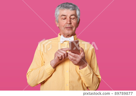 Stidio shot of mature white haired man dressed in yellow shirt and white bow tie, holding smartphone in hands and types message, looks concentrated on his gadget. People nd technology concept. 89789877