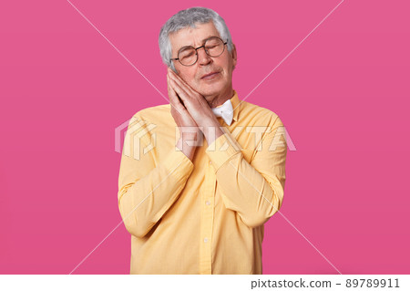 Sleepy grey haired mature man wears yellow shirt with bow tie poses with hands together while standing with closed eyes against rose wall. Male with short hairstyle wants to go to bad. People concept. 89789911