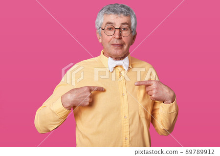 Closeup portrait of man in yellow shirt, asks question, poses in studio. Isolated over pink background. Tempered human emotions. Elderly male with glasses points with fore fingers on himself. 89789912