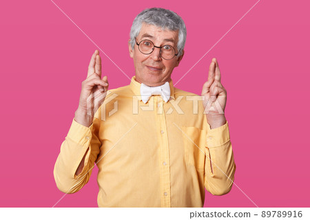 Studio shot of white haired senior raises hands up and crosses fingers. Elderly man wears yellow shirt poses in studio isolated over rose background, has hope for win lottery. People emotions concept. 89789916