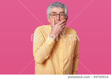 Closeup portrait of scared elderly man with short haistyle. Senior with opened mouth, keeps hand up to face, looks at camera with wide eyes, isolated on pink background. Facial expression. 89789919