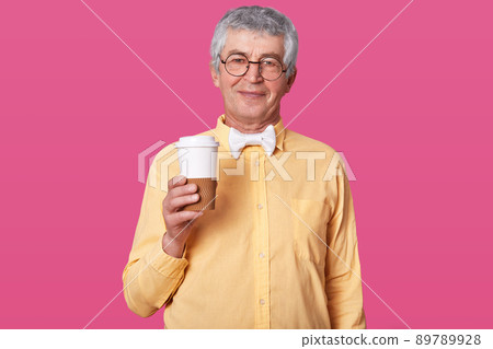 Elderly man stands with coffe against pink wall. Senior holds big cup ready for drinking hot baverage. Grey haired male with glasses and bowtie isolated over rose background. People concept. 89789928