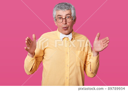 Astonished man shows large or wide object, wears modern bright shirt with white bowtie, has amazed facial expression. Pensioner shapes square, demonstrates size. Handsome old male against pink wall. 89789934