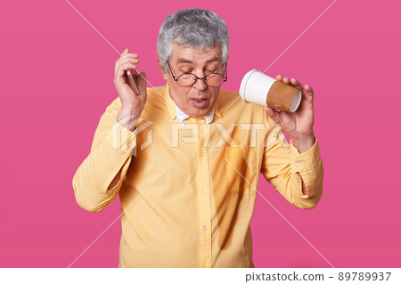 Close up portrait of eldery gray haired male poured takeaway coffee on his clothes while speaking on phone, has confused facial expression, poses isolated over pink studio background. Emotions concept 89789937