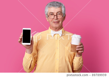 Photo of mature grey haired man in elegant shirt with bow tie, holds smart phone with mock up screen, takeaway coffee, looks directly at camera through round spectacles, isolated over pink studio wall 89789939
