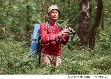 Excited male hitch hiker standing among trees with binoculars in forest, looks concentrated, eldery male wearing casual red sweater and hat, carries backpack and rug. Active recreation concept. 89790379