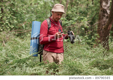 Close up portrait of concentrated eldery male wearing red casual sweater, pants and cap, standing in forest with compass and binoculars in hands, tries to find way, backpacking while having vacation. 89790385
