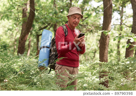 Experienced traveller holding his smartphone in one hand, using device for orientating, being alone, having compass and sleeping pad, wearing hat, trousers, sweatshirt, backpack. Travel concept. 89790398