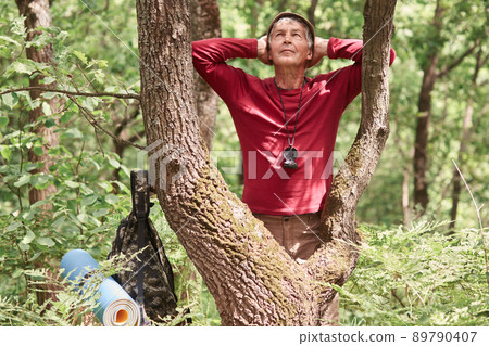 Hiker man hiking in forest, stops to have rest after long hours backpacking, healthy eldery sport person with backpack, stands near tree and enjoys wild nature, wears casually. Active rest concept. 89790407
