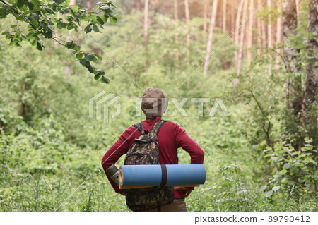 Hiking. Man wearing red sweater and cap in hike, having backpack and sleeping pad, looking towards. Sporty man hiking on green trees background. Rear view. Hiking, tourism, healthy lifeslyle concept. 89790412
