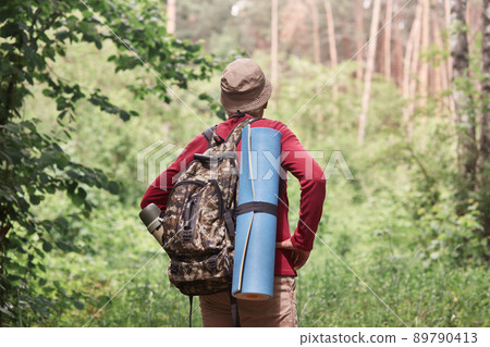 Back view of active backpacker looking for way out in forest, having sleeping pad and dark rucksack with thermos at his back, enjoying active holidays, stick to travel lifestyle. Travel concept. 89790413