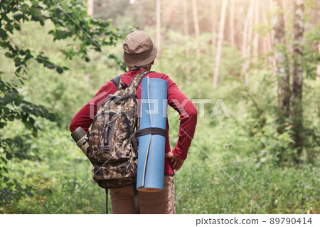 Outdoor shot of man with backpack and sleeping pad hiking in forest, forest landscape, back of tourist wears casual clothing and cap, standing with hands on hips and look far away. Lifestyle concept. 89790414