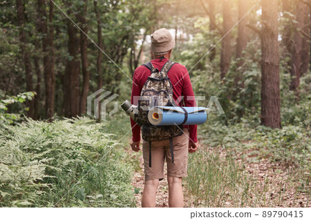 Backwards of man wearing hat, red sweater and beige trousers, posing with backpack and sleeping pad, looking at paths, surrounded with green trees. Traveling, lifestyle and adventure concept. 89790415