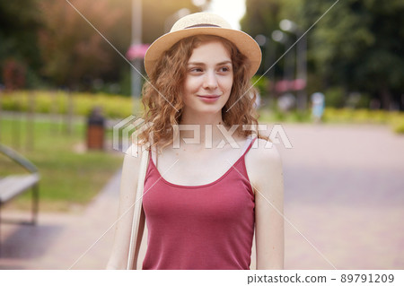 Romantic girl wearing straw hat and burgundy t shirt posing in park, looking smiling aside, enjoing beautiful nature. Adorable wavy haired womaning walk in park, waiting her friend, having holiday. 89791209