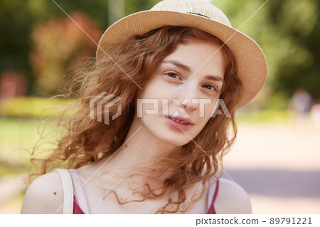 Close up portrait of attractive young lady in straw hat. Foxy haired charming woman with brown eyes looking at camera with gentle, has calm facial expression, posing isolated over park background. 89791221