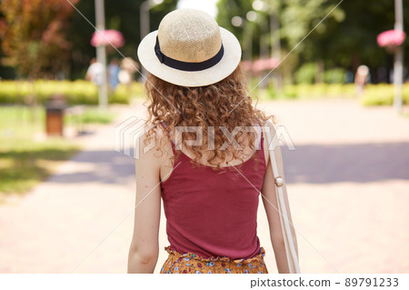 Back view of slender curly haired female wearing straw hat and red shirt, having bag on her shoulder, walking along paved street, spending time around nature in local park. Summer day concept. 89791233