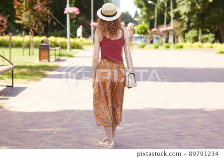 Full length photo from back view of young slender lady walking in tree shadow, fond of nature, spending lunchtime in recreation zone, wearing red shirt, orange skirt, sshoulder bag and straw hat. Full length photo from back view of young slender lady walking in tree shadow, fond of nature, spending lunchtime in recreation zone, wearing red shirt, orange skirt, sshoulder bag and straw hat. 89791234