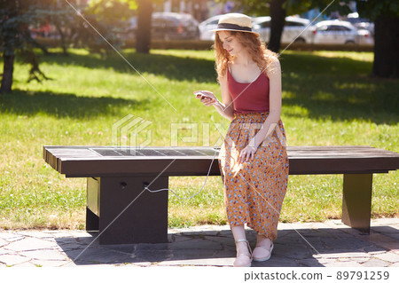 Image of young girl charges mobile phone via USB outdoors, female sitting on bench with solar panel in town park. Public charging, modern technology, alternative electricity, renewable energy concept. 89791259