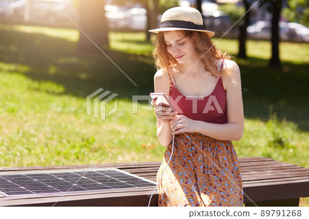 Horizontal short of young girl charges mobile phone via USB outdoors, lady sits on bench with solar panel in town park. Public charging on city street. Modern technology, ecology, alternative energy. 89791268