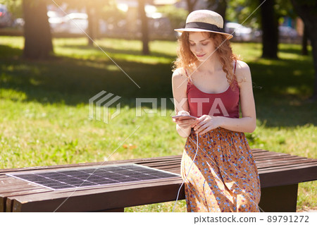 Beautiful woman charging her phone on free multipurpose solar panel, attractive female sitting on bench intown park. Modern technology, free energy for everyone, solar panel, ecology concept. Beautiful woman charging her phone on free multipurpose solar panel, attractive female sitting on bench intown park. Modern technology, free energy for everyone, solar panel, ecology concept. 89791272